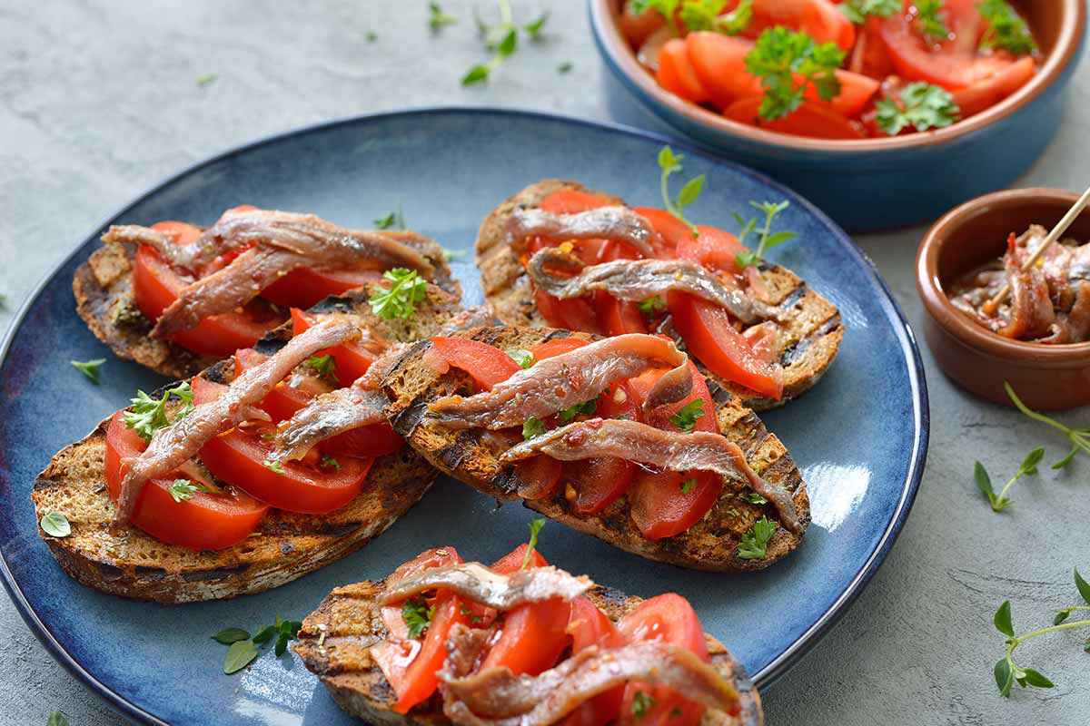 Piatto celeste con sopra del pane abbrustolino con pomodori rossi maturi e acciughe sotto sale