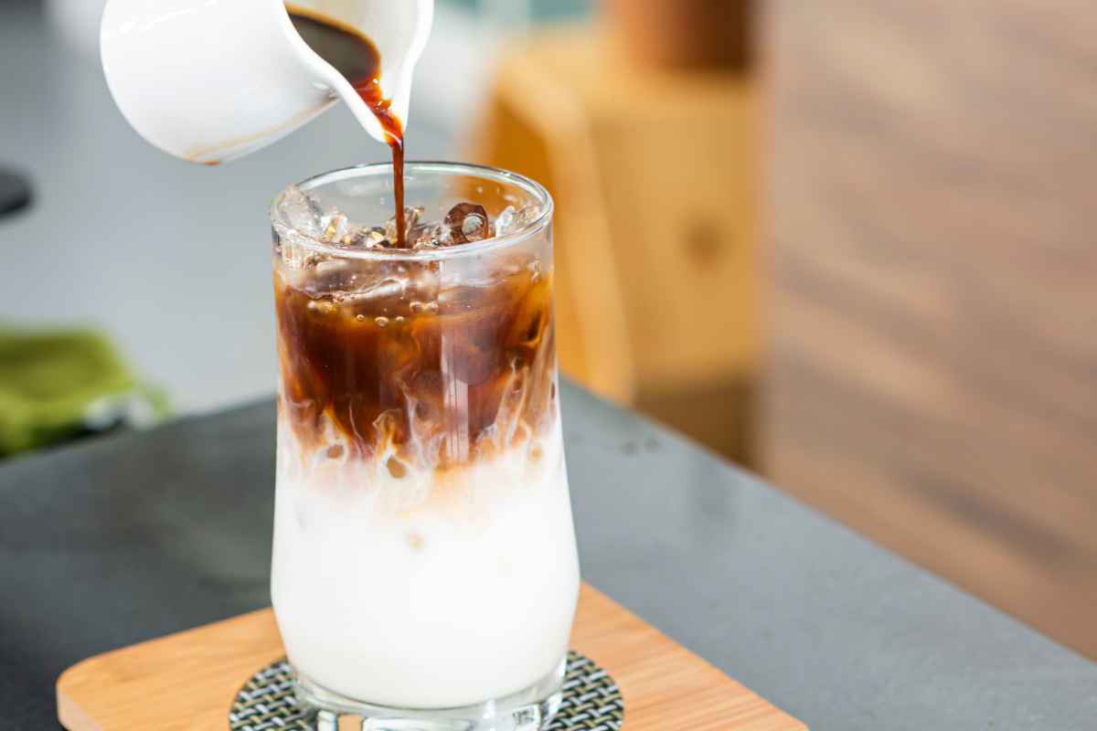 bicchiere di vetro alto con latte di mandorla, ghiaccio e caffè versato sopra a filo