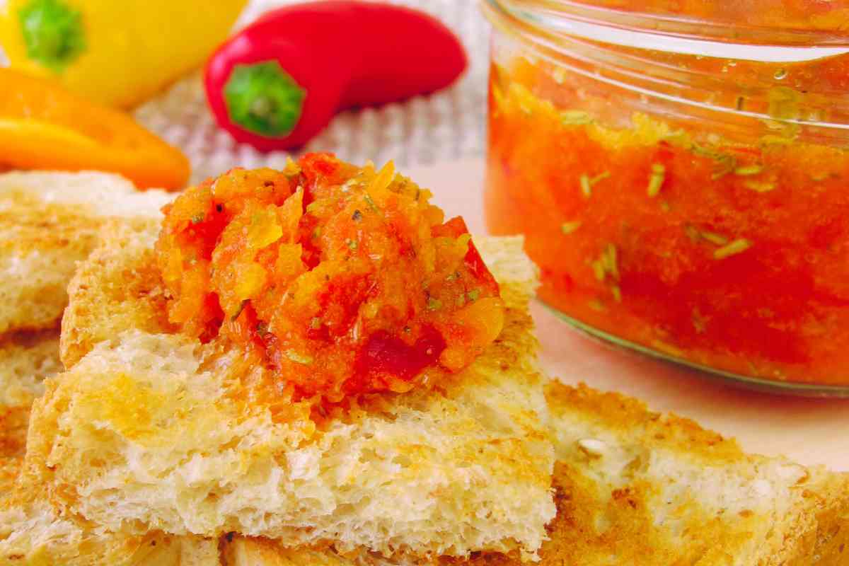 bruschetta di pane casereccio con crema di peperoni vicino a un vasetto pieno di crema e peperoni sullo sfondo