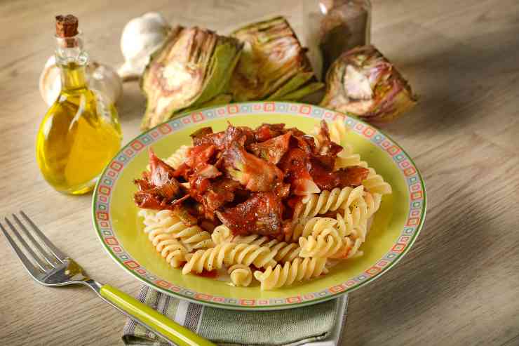 Pasta con carciofi al sugo di pomodoro in un piatto con dietro carciofi a spicchi, aglio e un'oliera