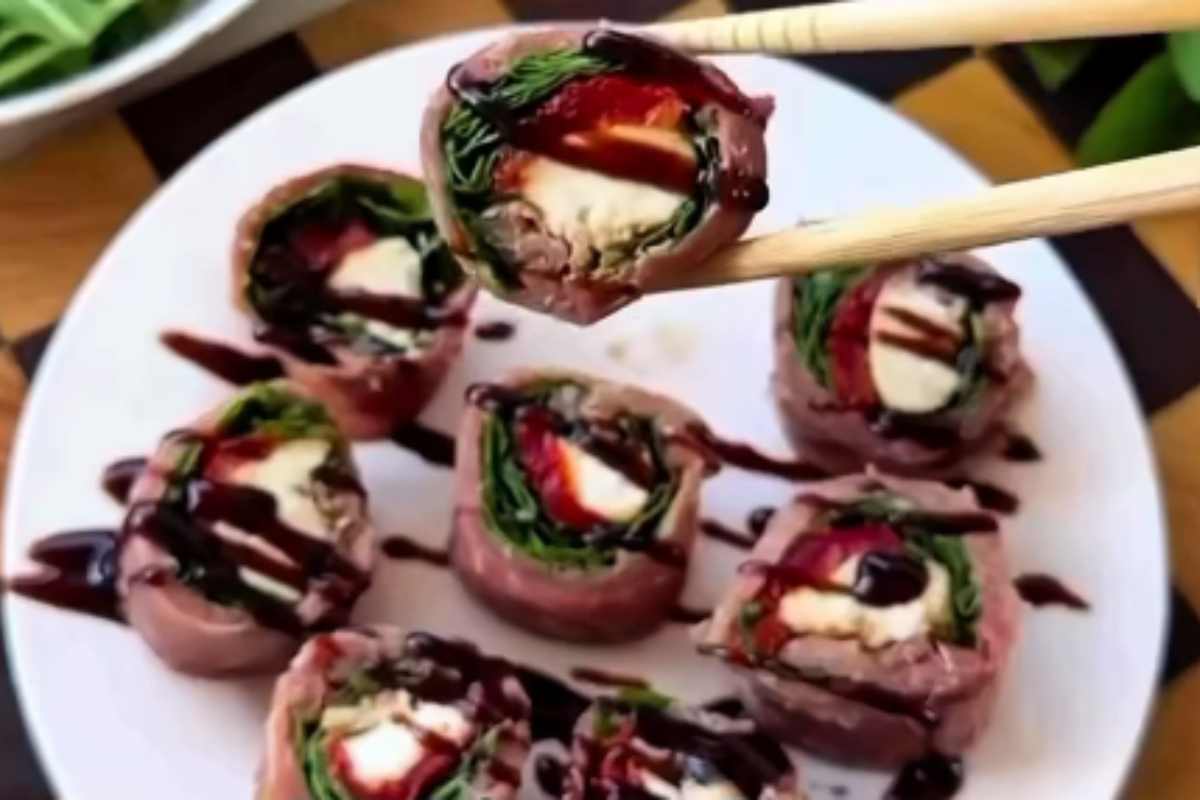 Involtini di prosciutto crudo mozzarella pomodori secchi e rucola in un piatto con un rotolino sospeso tra bacchette di legno