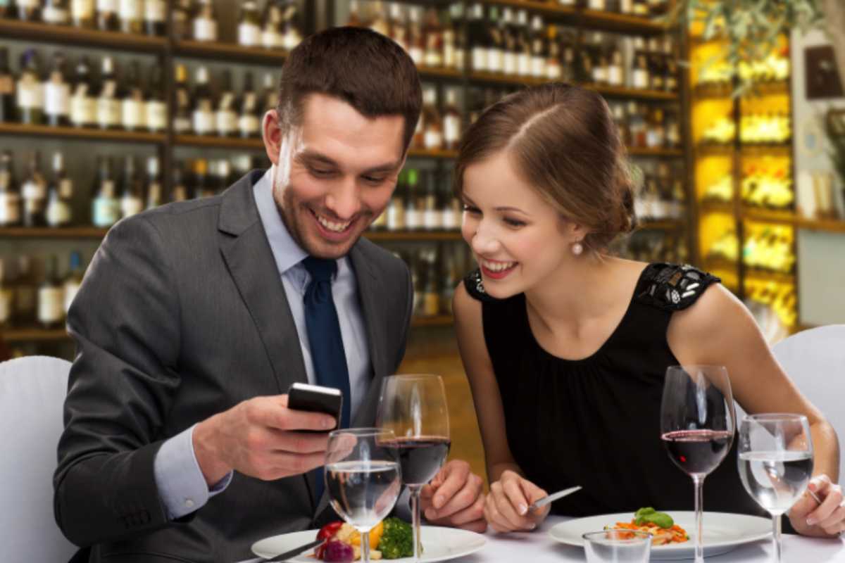 uomo e donna seduti al tavolo di un ristorante che sorridono mentre guardano qualcosa sul cellulare