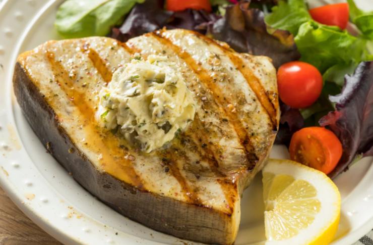 trancio di pesce spada con sopra una salsa e accanto insalata e pomodori