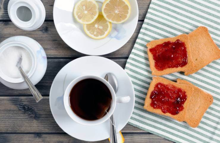 fette biscottate con marmellata e accanto una tazza di tè e dello zucchero