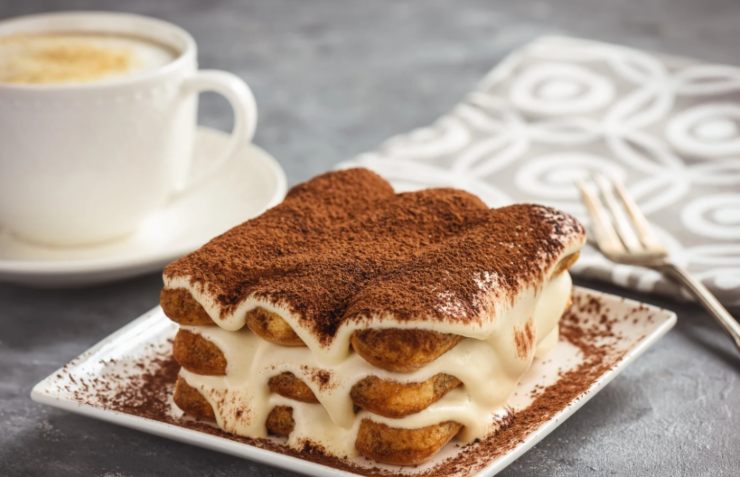 fetta di tiramisù su un piattino e dietro un cappuccino