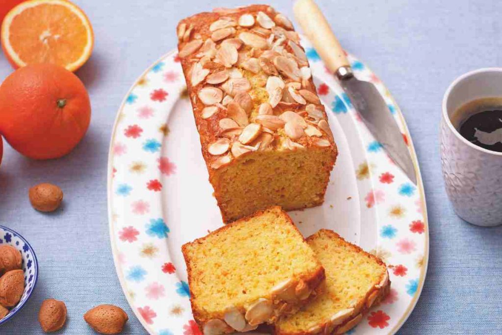 vassoio di plumcake all'arancia su un tavolo con vicino mandorle, arance e una tazza di caffè