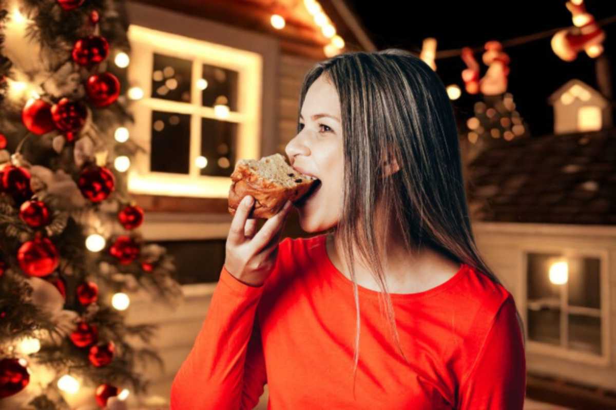 donna che mangia una fetta di panettone e accanto l'albero di natale illuminato