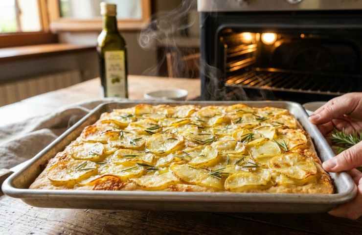 Focaccia di patate e rosmarino appena sfornata, ancora fumante nella teglia di metallo, estratta dal forno acceso in una cucina domestica.