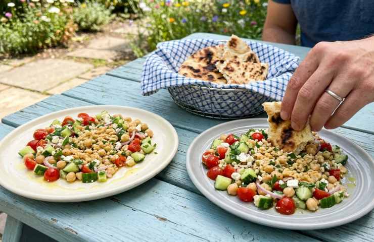 Insalata di ceci schiacciati servita con pane pita caldo su un tavolo all'aperto, ricetta sana estiva con verdure fresche e feta greca in due piatti.