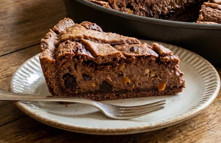 Fetta di pastiera napoletana al cioccolato su piatto, con tortiera sullo sfondo, su tavolo di legno.