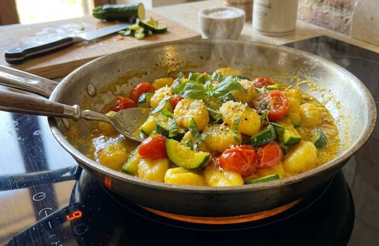 Gnocchi allo zafferano con zucchine e pomodorini in fase di cottura e mantecatura in una padella di acciaio sul piano cottura a induzione. Vista dall'alto della preparazione cremosa senza panna.