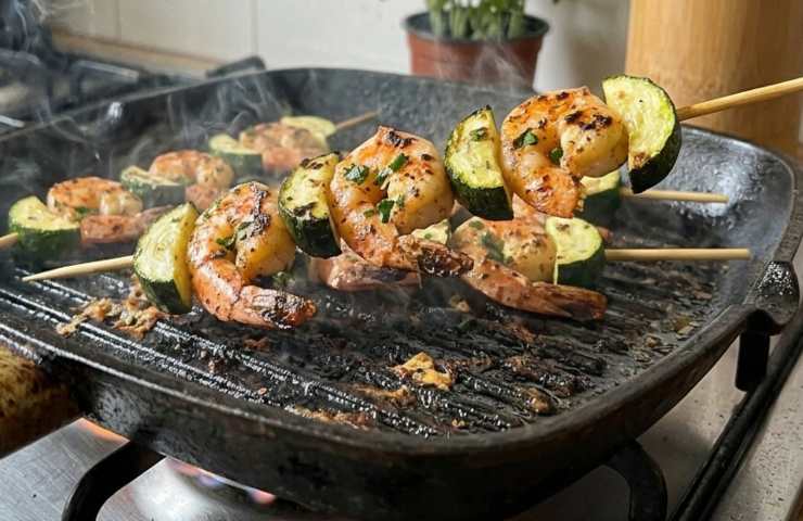 Spiedini di gamberi e zucchine mentre cuociono su una piastra in ghisa rovente sul fornello di casa, con fumo leggero e segni di grigliatura.
