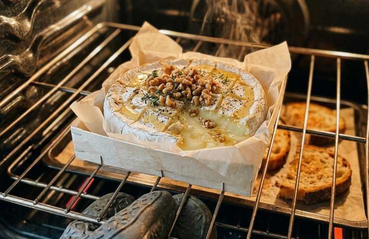 Forma di Camembert che si scioglie in forno sopra una griglia metallica, con gherigli di noci e miele in superficie e fette di pane casereccio che tostano accanto.