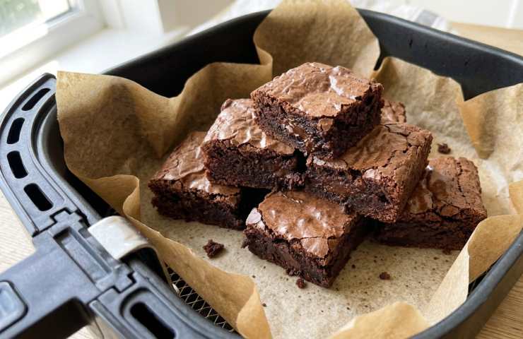 Brownies al cioccolato appena cotti su carta forno all'interno del cestello di una friggitrice ad aria nera, posizionata su un tavolo di legno.