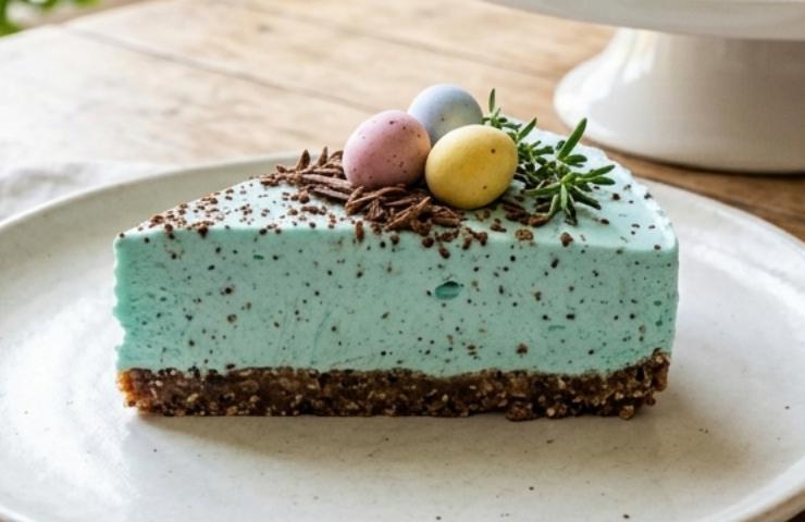 Fetta di cheesecake di Pasqua color Tiffany con ovetti confettati su piatto bianco e tavolo in legno luminoso.