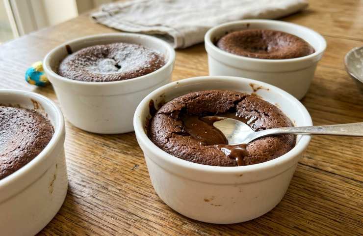Foto realistica di quattro tortini dal cuore morbido al cioccolato delle uova di Pasqua in pirottini bianchi su tavolo in legno, con cucchiaio che rivela il centro colante.