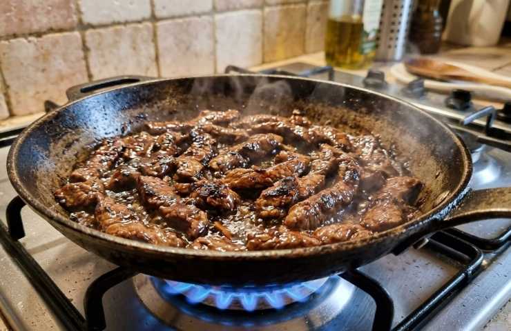 Straccetti di manzo in padella che sfrigolano sul fornello a gas durante la sfumata con aceto balsamico.
