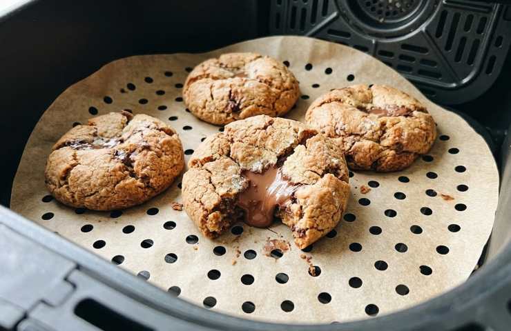 Primo piano di cookies alla Nutella appena cotti in friggitrice ad aria, posizionati su un foglio di carta forno bucherellata all'interno del cestello. Un biscotto spezzato mostra il cuore cremoso e fuso.