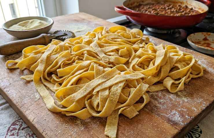 Primo piano di tagliatelle fresche all'uovo fatte a mano, infarinate con semola su un tagliere di legno, con una pentola di ragù di carne e una ciotola di farina in background in una cucina luminosa.
