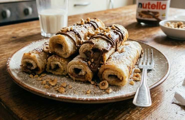 Vassoio di rotolini di pancarré alla Nutella con zucchero a velo e granella di nocciole, serviti a colazione su un tavolo in legno con un bicchiere di latte.