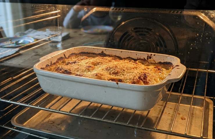 Teglia di ceramica bianca con sformato di cavolfiore cremoso in cottura nel forno ventilato, superficie gratinata con parmigiano e pangrattato che bolle, vista attraverso il vetro del forno acceso.