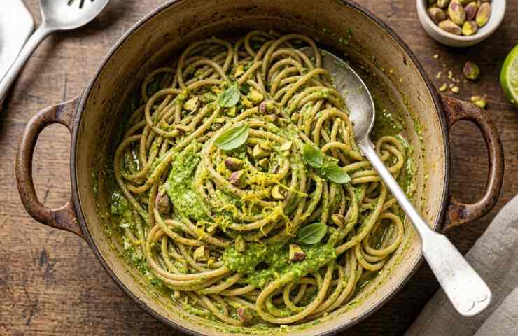 Inquadratura dall'alto di una pentola in ghisa riempita con spaghetti integrali appena saltati e cremosi, conditi con pesto di pistacchi verde brillante, basilico fresco, granella e scorza di lime, pronti da servire.