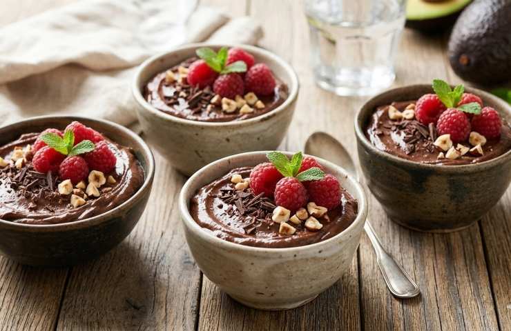 Quattro coppette monoporzione di mousse di avocado e cacao disposte su un tavolo di legno, decorate con lamponi freschi, granella di nocciole e foglioline di menta, pronte da servire.