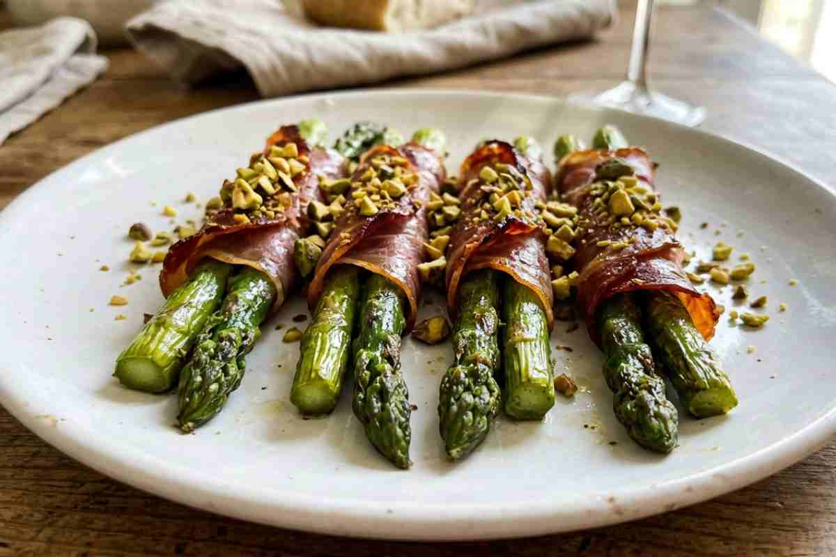 Asparagi avvolti in prosciutto crudo e granella di pistacchio su piatto bianco.