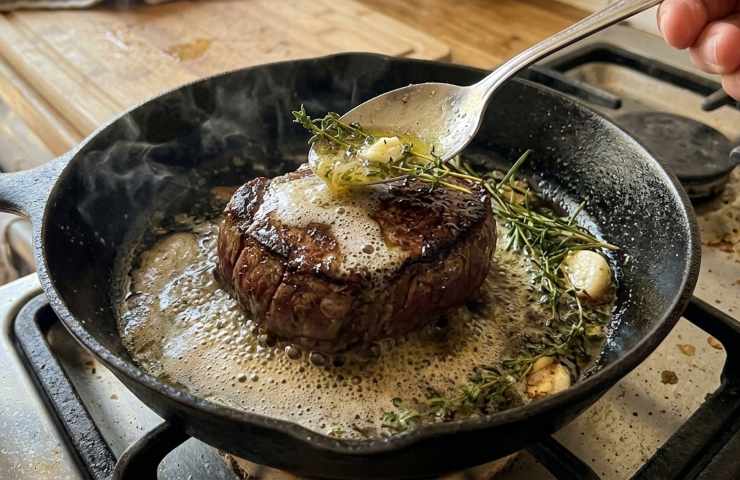 Inquadratura ravvicinata della tecnica arrosé: un cucchiaio versa burro fuso spumeggiante, aglio e rosmarino su un filetto di manzo in una padella di ghisa fumante.