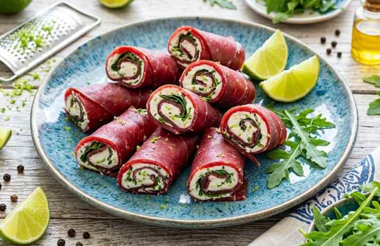 Involtini di bresaola con ripieno cremoso al caprino e rucola, serviti con spicchi di lime fresco su un piatto blu decorato.