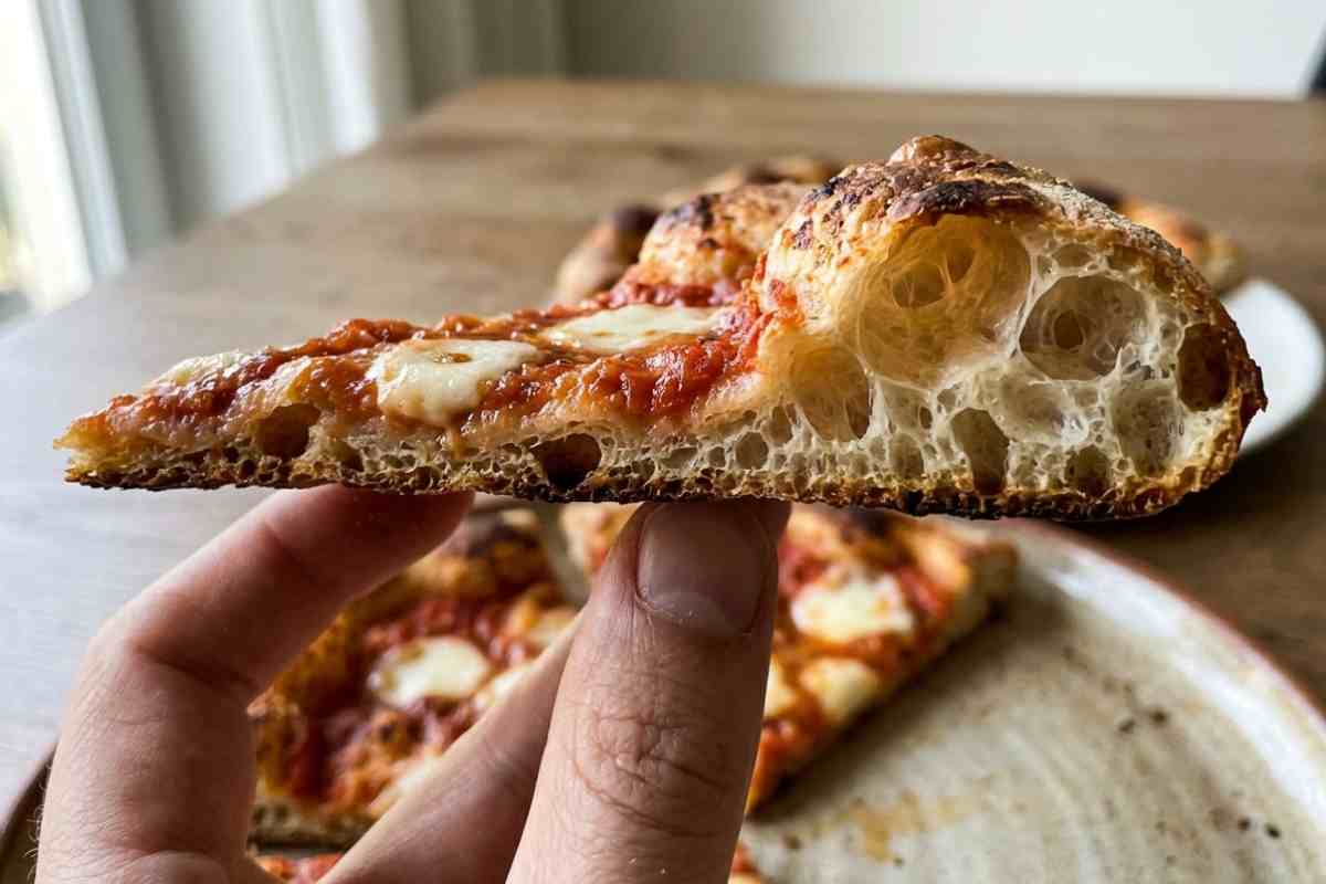 Fetta di pizza scrocchiarella sollevata di profilo che mostra un'alveolatura estrema con grandi bolle d'aria e base croccante.