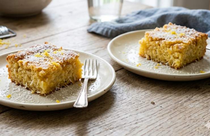 Due quadrotti soffici al limone e cocco serviti su piatti da dessert in ceramica, con spolverata di zucchero a velo e scorza gialla su un tavolo in legno luminoso.