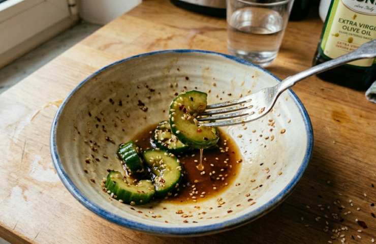 Piatto di cetrioli smashed quasi finito servito con forchetta all'interno su tavolo con bicchiere d'acqua.
