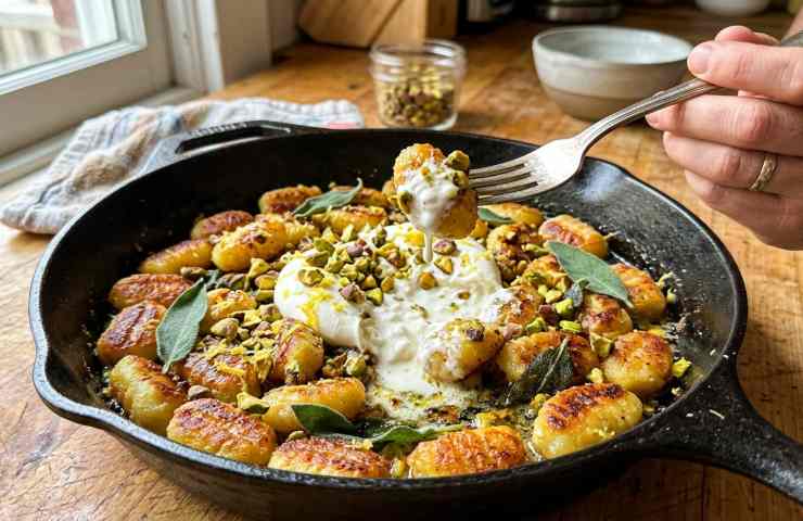 Gnocchi di patate croccanti in padella con burrata fresca, granella di pistacchi e foglie di salvia, serviti in una padella di ghisa su un tavolo di legno.