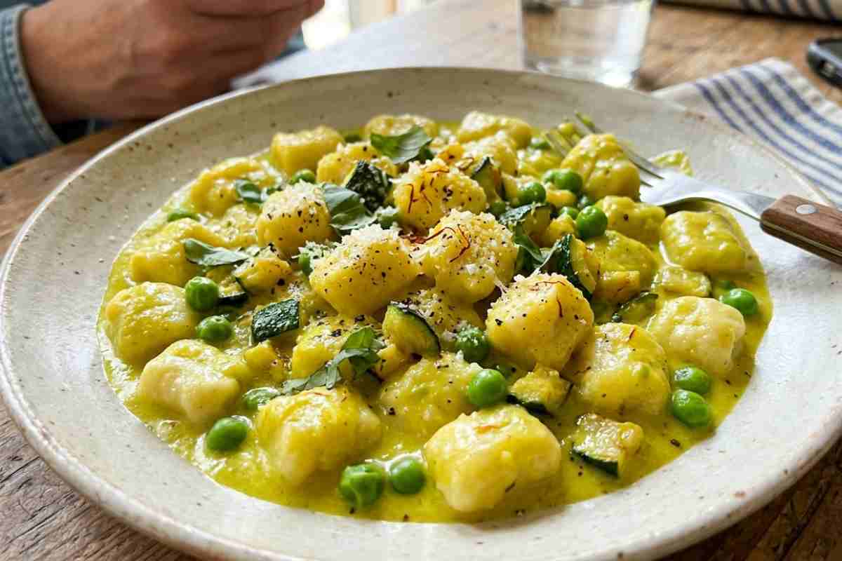 Piatto di gnocchi di ricotta freschi alla crema di zafferano, piselli e zucchine su tavola di legno, con una forchetta.